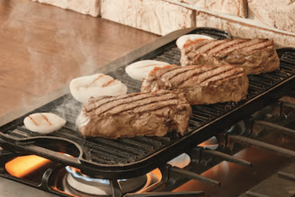 Cortes de carne dorándose a la plancha sobre una parrilla acanalada en una cocina de gas.
